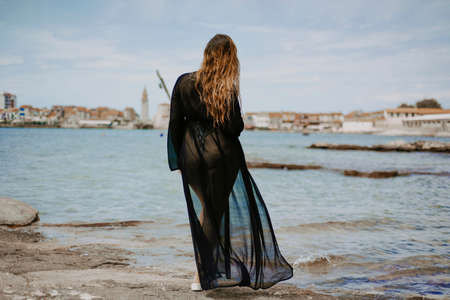 Gorgeous brunette on the beach against the backdrop of the sea and the European city. Woman with long hair and flowing dressの写真素材