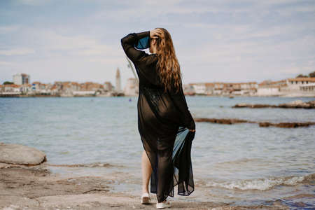 Gorgeous brunette on the beach against the backdrop of the sea and the European city. Woman with long hair and flowing dressの写真素材