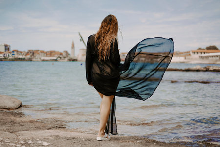 A woman with a flying pareo. Gorgeous brunette on the beach against the backdrop of the sea and the European city.の写真素材