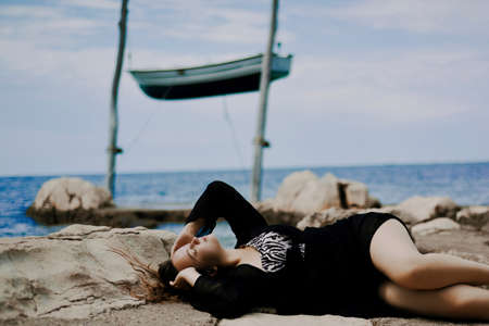 Gorgeous brunette on the beach against the backdrop of the sea and a suspended boat. Woman with long hair resting, summer atmospheric photo.の写真素材