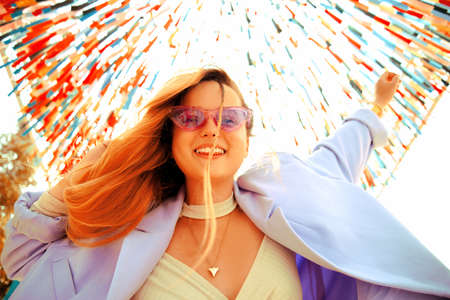 A girl at the festival looks at the camera and smiles against the background of a colorful background, dancing in purple glasses and a jacket with long hairの写真素材