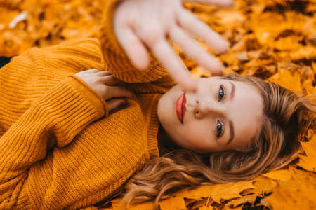 Beautiful girl lies in autumn leaves in the park, autumn. Young girl closes the camera with her handの写真素材
