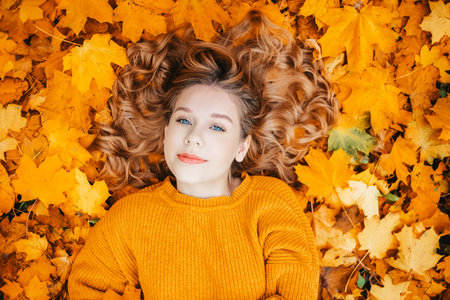 A beautiful young girl with blue eyes lies in the autumn leaves in the park and looks into the camera, smilesの写真素材