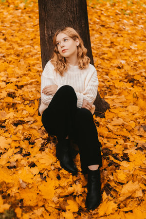 A girl in a white sweater sits on the autumn leaves. A beautiful blonde with a square looks at the camera. Woman outdoors in the autumn parkの写真素材