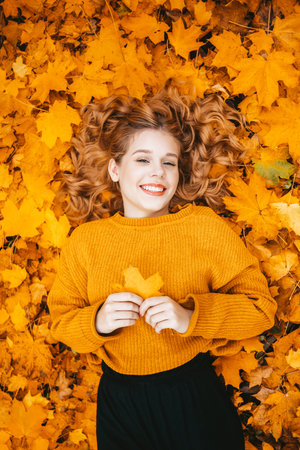 A beautiful girl lies in autumn leaves in the park, gorgeous hair, a teenager smiles and looks at the camera in autumnの写真素材