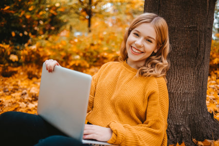 A beautiful girl teenager sits in an autumn park with a laptop. The student studies on the street. Study in the fall. The woman looks into the camera.の写真素材