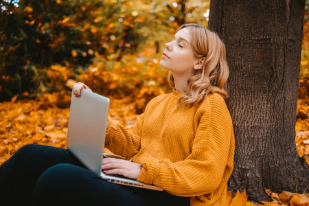 A beautiful girl teenager sits in an autumn park with a laptop. The student studies on the street. The girl closed her eyes and restedの写真素材