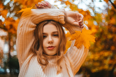 Photo portrait of a girl in a white sweater, the blonde holds leaves in her hands. Pretty woman in the park in autumn. Autumn fashion and style. Warm moodの写真素材