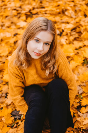 A girl in a white sweater sits on the autumn leaves. A beautiful blonde with a square looks at the camera. Woman outdoors in the autumn park.の写真素材