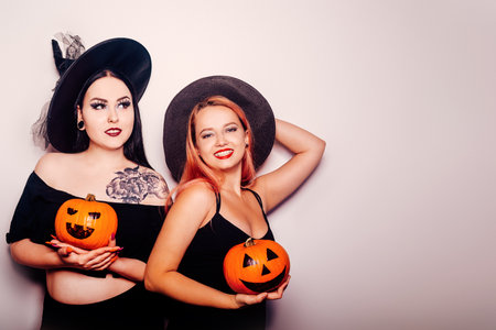Two girls in witch costumes on a white background hold pumpkins, smile and laugh for Halloween. Autumn photo of girlfriends from a costume partyの写真素材