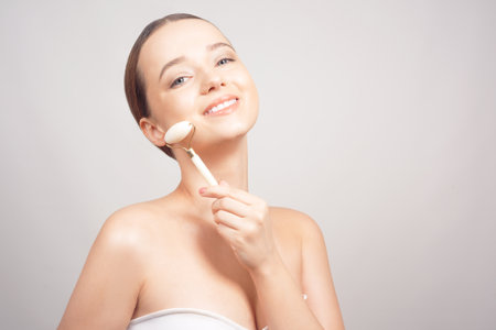 Beauty face care. Woman doing face massage with white jade facial rollers for spa skin care treatment at home. Girl model using natural massager tool portrait on whiteの写真素材