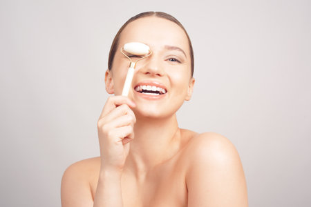 Beauty face care. Woman doing face massage with white jade facial rollers for spa skin care treatment at home. Girl model using natural massager tool portrait on whiteの写真素材