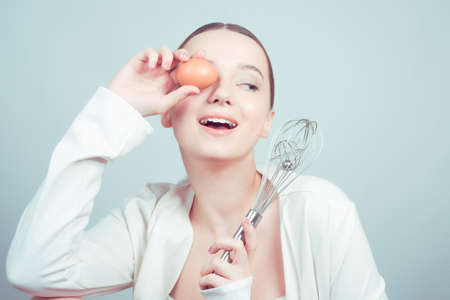 The girl stands in white background, balloon whisk and aggs and cooks, cooking courses, school cooks. The girl grimaces, shows emotions, is surprised, smilesの写真素材