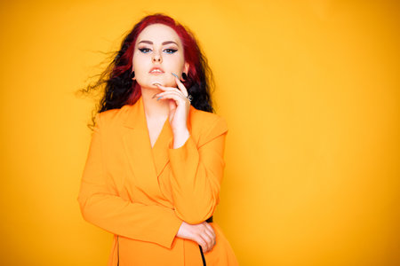 Beautiful girl on a yellow background. A brunette in an orange dress and long hair is posing, smiling, having fun. Summer photo woman in studio. Joy and emotion. Red hair roots place for textの写真素材