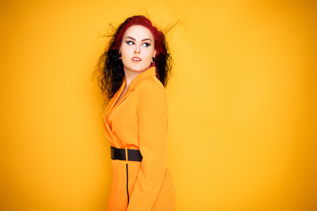 Beautiful girl on a yellow background. A brunette in an orange dress and long hair is posing, smiling, having fun. Summer photo woman in studio. Joy and emotion. Red hair roots place for textの写真素材