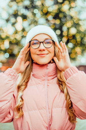 Cute portrait of a girl in glasses on the background of a Christmas tree. Winter, the first snow, a blonde in a pink jacket and a white hat smiles. New Year.の写真素材