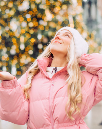 A cute blonde in a hat and a pink jacket against the background of a Christmas tree on the street. Winter walk, new year, feast dayの写真素材