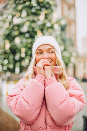 Cute portrait of a girl in the winter on the street, the snow is falling, the blonde, Christmas tree, warms her hands. Girl in a white hat, pink jacket on the background of a tree, new yearの写真素材