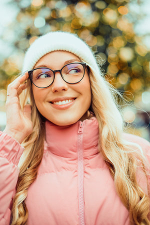Cute portrait of a girl in glasses on the background of a Christmas tree. Winter, the first snow, a blonde in a pink jacket and a white hat smiles. New Year.の写真素材