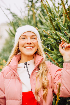 A cute blonde chooses a Christmas tree for the new year. A woman in a hat and pink jacket smiles at the camera in snowy weather. Winter holidays and new yearの写真素材