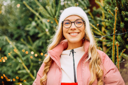 A cute blonde chooses a Christmas tree for the new year. A woman in a hat and pink jacket smiles at the camera in snowy weather. Winter holidays and new yearの写真素材