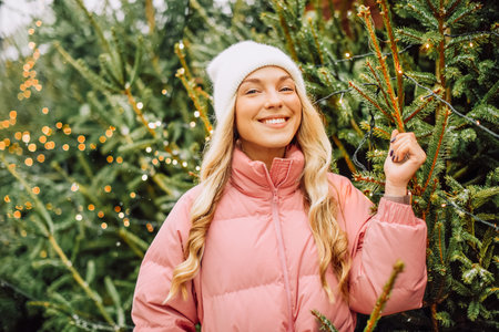 A cute blonde chooses a Christmas tree for the new year. A woman in a hat and pink jacket smiles at the camera in snowy weather. Winter holidays and new yearの写真素材