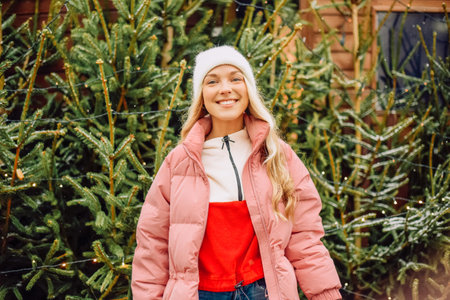 A cute blonde chooses a Christmas tree for the new year. A woman in a hat and pink jacket smiles at the camera in snowy weather. Winter holidays and new yearの写真素材