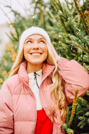 A cute blonde chooses a Christmas tree for the new year. A woman in a hat and pink jacket smiles at the camera in snowy weather. Winter holidays and new yearの写真素材