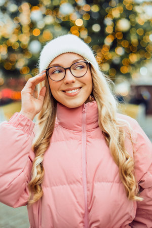 Cute portrait of a girl in glasses on the background of a Christmas tree. Winter, the first snow, a blonde in a pink jacket and a white hat smiles. New Year.の写真素材