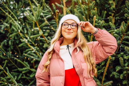 A cute blonde in a hat and a pink jacket and glasses stands against the background of Christmas trees. The girl chooses a Christmas tree, New Year's moodの写真素材
