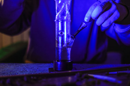 Male hands clog a bong with marijuana. Drugs and drug addict. The man smokes weed. Neon blue light....