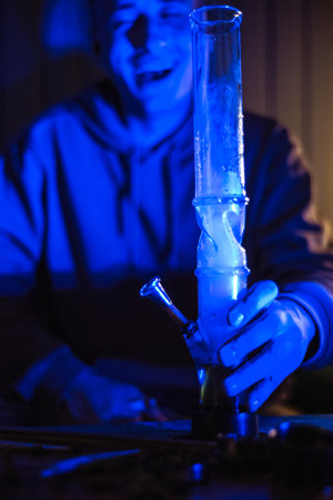 Male hands clog a bong with marijuana. Drugs and drug addict. The man smokes weed. Neon blue light. CBD or THC. Man laughs, arrival, relaxationの写真素材