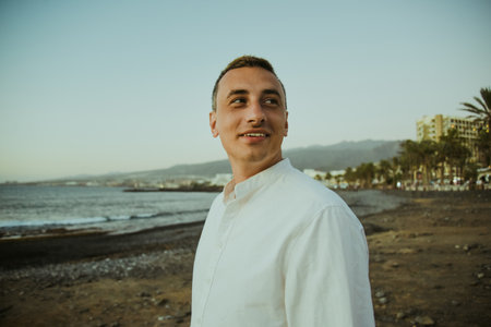 A man in a white shirt against the background of the sea and sunset. A guy is walking on the beach and enjoying the views, happiness from traveling in the summer, freedom.の写真素材