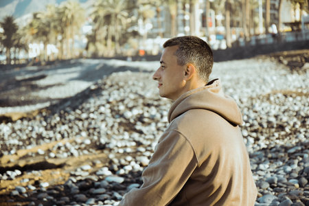 A man in a beige hoodie sits on the background of the sea. A guy is walking on the beach and enjoying the views, happiness from traveling in the summer, freedom. yoga, meditationの写真素材