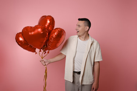 A man is holding red balloons in the shape of a heart on a pink background. Valentine's Day, love, February 14. Looks at the balloonsの写真素材