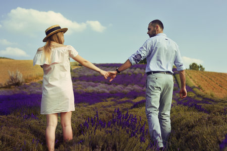 A loving couple is walking along the lavender field, a girl in a dress and a straw hat with a man holds hands and walks through the field. Romantic relationship in a couple. Beauty, unity with nature.の写真素材