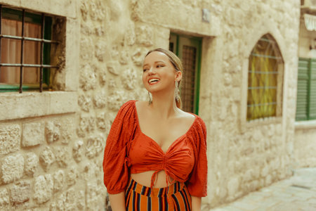 Beautiful stylish girl with long earrings, wearing an orange suit looks at the camera. Traveling around Europe Fashionable woman smiling in the old town and posing.の写真素材