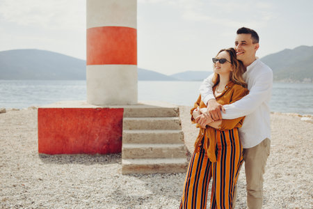 A couple stands near a lighthouse on the beach against the backdrop of the sea and mountains. Vacation love, honeymoon, travel. Stylish couple laughing and smilingの写真素材
