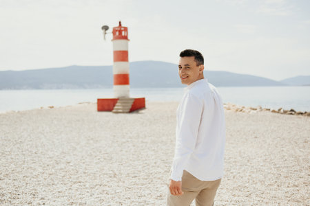 A man in a white shirt walks along the beach. The guy against the backdrop of mountains and the sea goes to the red lighthouse. Rest and travelの写真素材