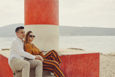 A couple sits near a lighthouse on the beach against the backdrop of the sea and mountains. Vacation love, honeymoon, travel. stylish coupleの写真素材