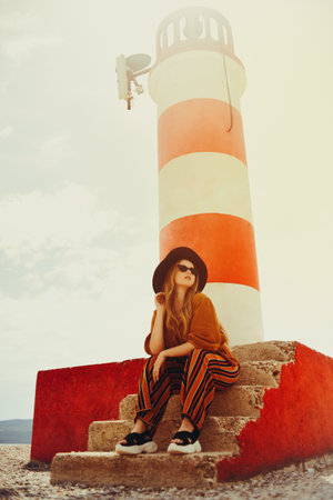 Gorgeous fashionable woman posing while sitting on a lighthouse. Summer photo of a girl in a yellow-orange suit, black hat and sunglasses. Blonde girl posing in travelの写真素材