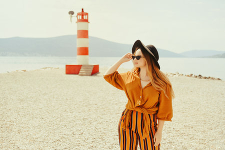 A girl in a yellow-orange suit on the beach against the background of the sea, mountains and a lighthouse. Fashion woman in sunglasses and hat dancingの写真素材