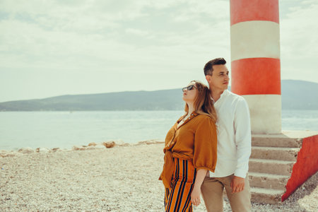 A couple stands near a lighthouse on the beach against the backdrop of the sea and mountains. Vacation love, honeymoon, travel. Stylish couple laughing and smilingの写真素材