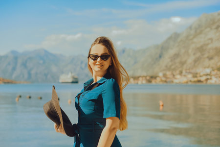 A woman in sun glasses and a hat stands against the backdrop of the sea and mountains. vacation and outdoor recreation. Girl with long hair on vacationの写真素材