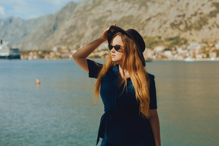 A woman in sun glasses and a hat stands against the backdrop of the sea and mountains. vacation and outdoor recreation. Girl with long hair on vacationの写真素材