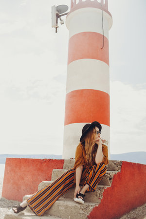 Gorgeous fashionable woman posing while sitting on a lighthouse. Summer photo of a girl in a yellow-orange suit, black hat and sunglasses. Blonde girl posing in travelの写真素材