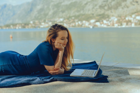 Girl freelancer lies on the background of the mountains with a laptop, remote work. A woman looks at the screen and writes something on the beach in the summer.の写真素材