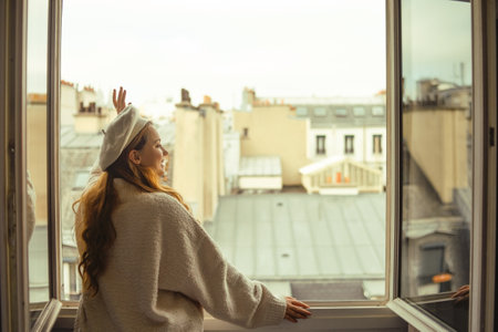 A woman stands near the window against the backdrop of the roofs of Paris. A girl with long hair in a beige suit and a beret enjoys the city and the trip waving to her neighbors.の写真素材