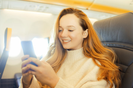 A beautiful girl is sitting in an airplane looking at the phone, reading a book or playing games. Air travel, long flight.の写真素材