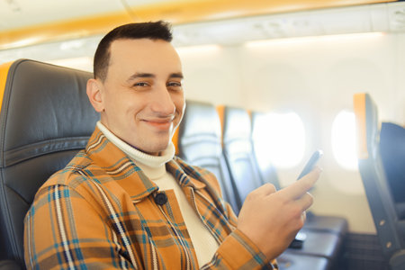 A man in an airplane looks at the phone, Wi-Fi in the airplane cabin. A tourist in transport in the sky uses a smartphone. The man is looking at the cameraの写真素材
